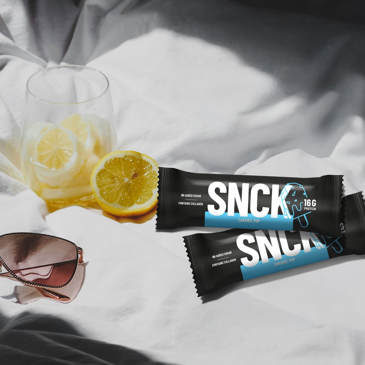 Two SNCK protein bars on a white surface with lemons and a glass of lemonade.