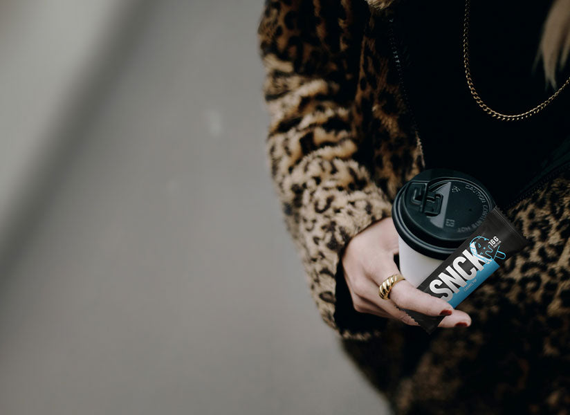 Person holding a coffee cup with a visible SNCK Bar, wearing a leopard print coat.